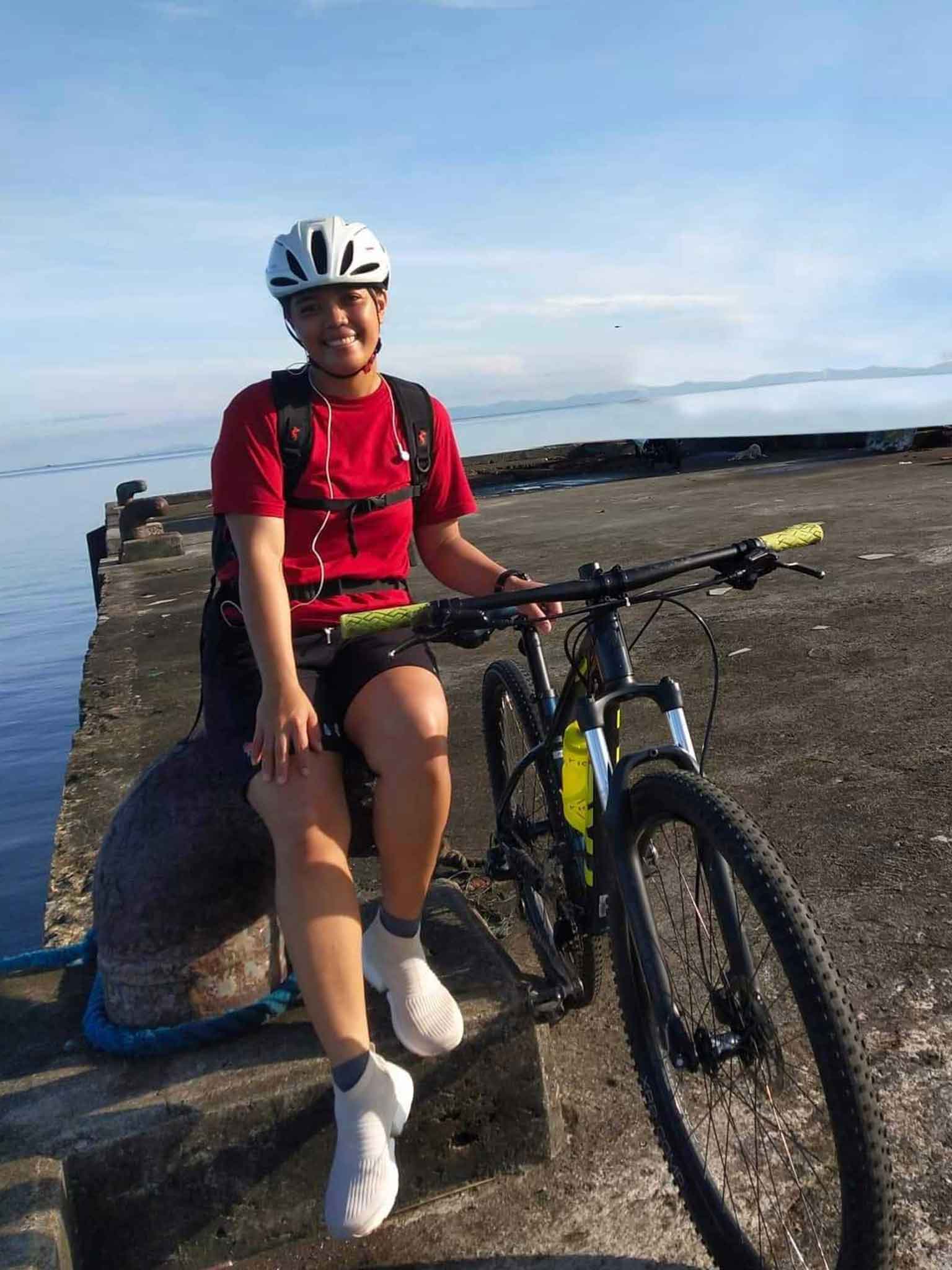 AB Cherry Opena, a female seafarer, sits on a pier with her bike, wearing a red cycling outfit and helmet, near the sea.