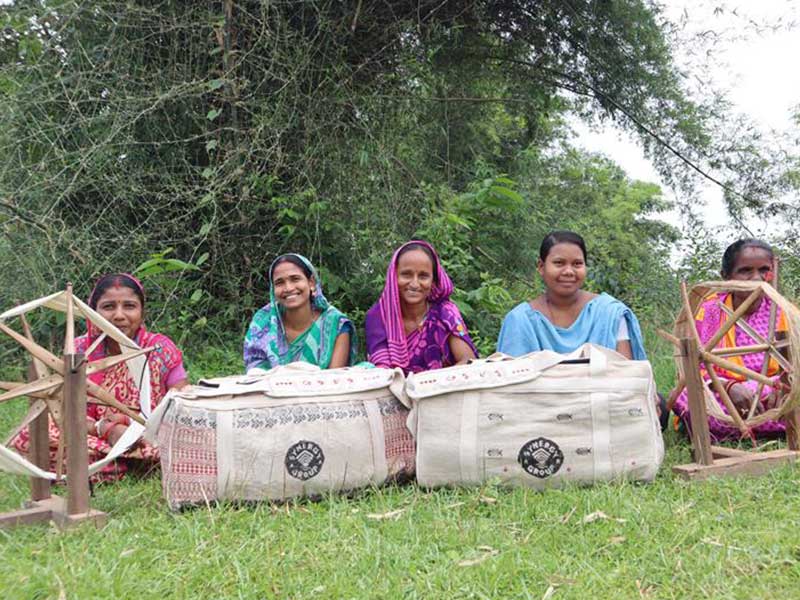 Five tribal women in colorful sarees seated outdoors with handloom tools and Synergy Marine Group juco bags in front of them