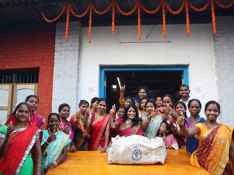 A group of women weavers and community members cheerfully pose with a Synergy Marine Group juco bag outside their centre