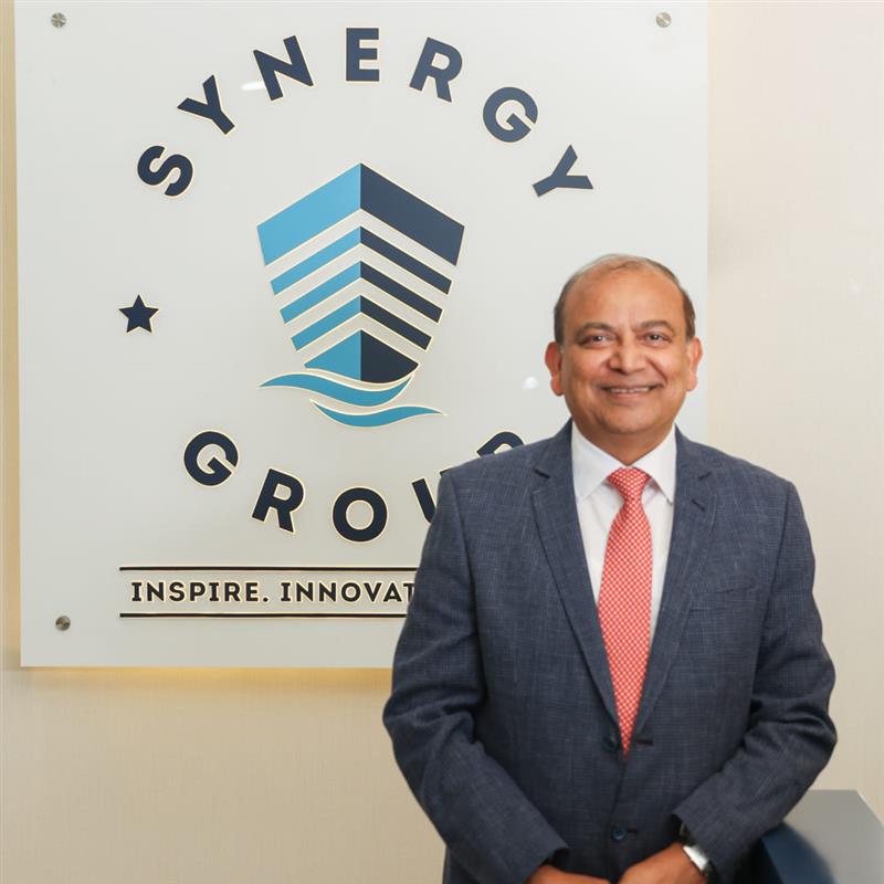 Ajay Chaudhry, Co-CEO – Ship Management, at Synergy Marine Group, photographed in front of a wall featuring the company logo.