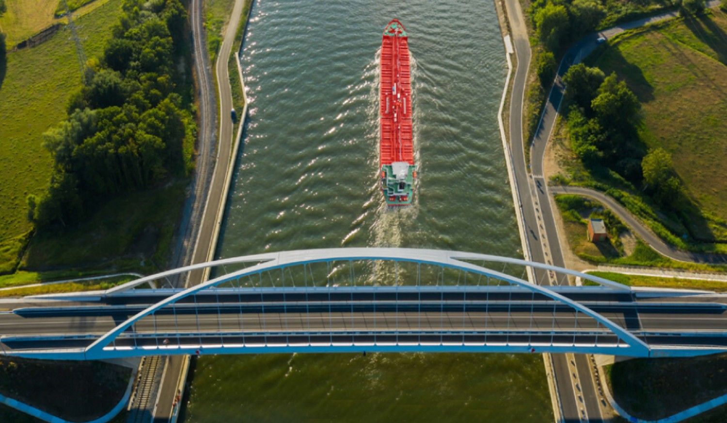 High-angle aerial view of a red cargo ship passing under a modern white arch bridge, symbolizing the integration of maritime trade with sustainable environments.