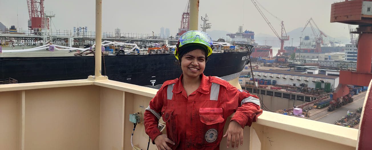 Fourth Engineer at Synergy Marine Group, standing on deck in PPE with a shipyard and vessel in the background, representing her role in marine engineering and ship operations.