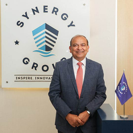 Ajay Chaudhry, Co-CEO of Synergy Marine Group, at the Synergy office reception with company logo.