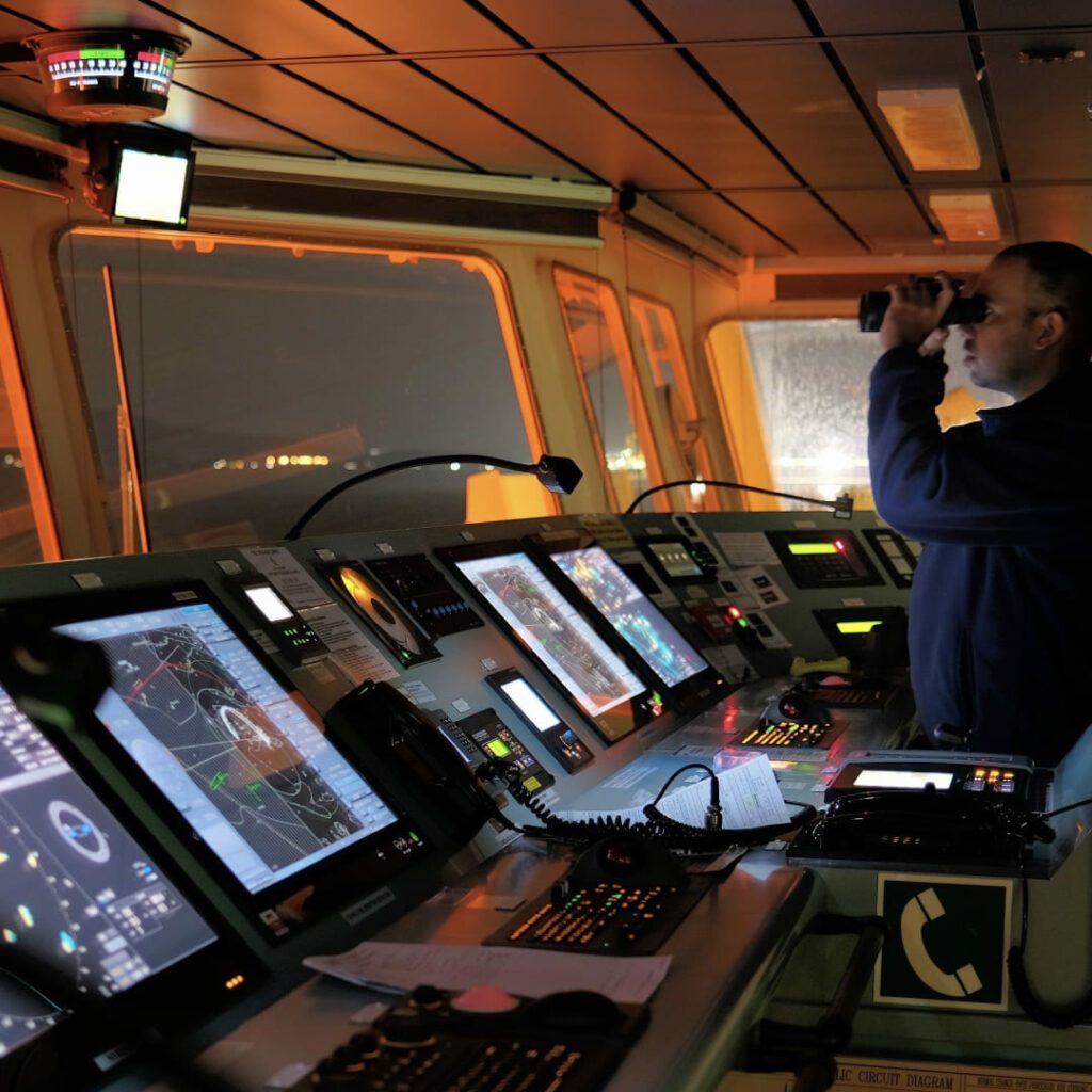 A detailed view of a ship’s bridge navigation console at night, featuring illuminated electronic displays and control panels, with a crew member using binoculars in the background.