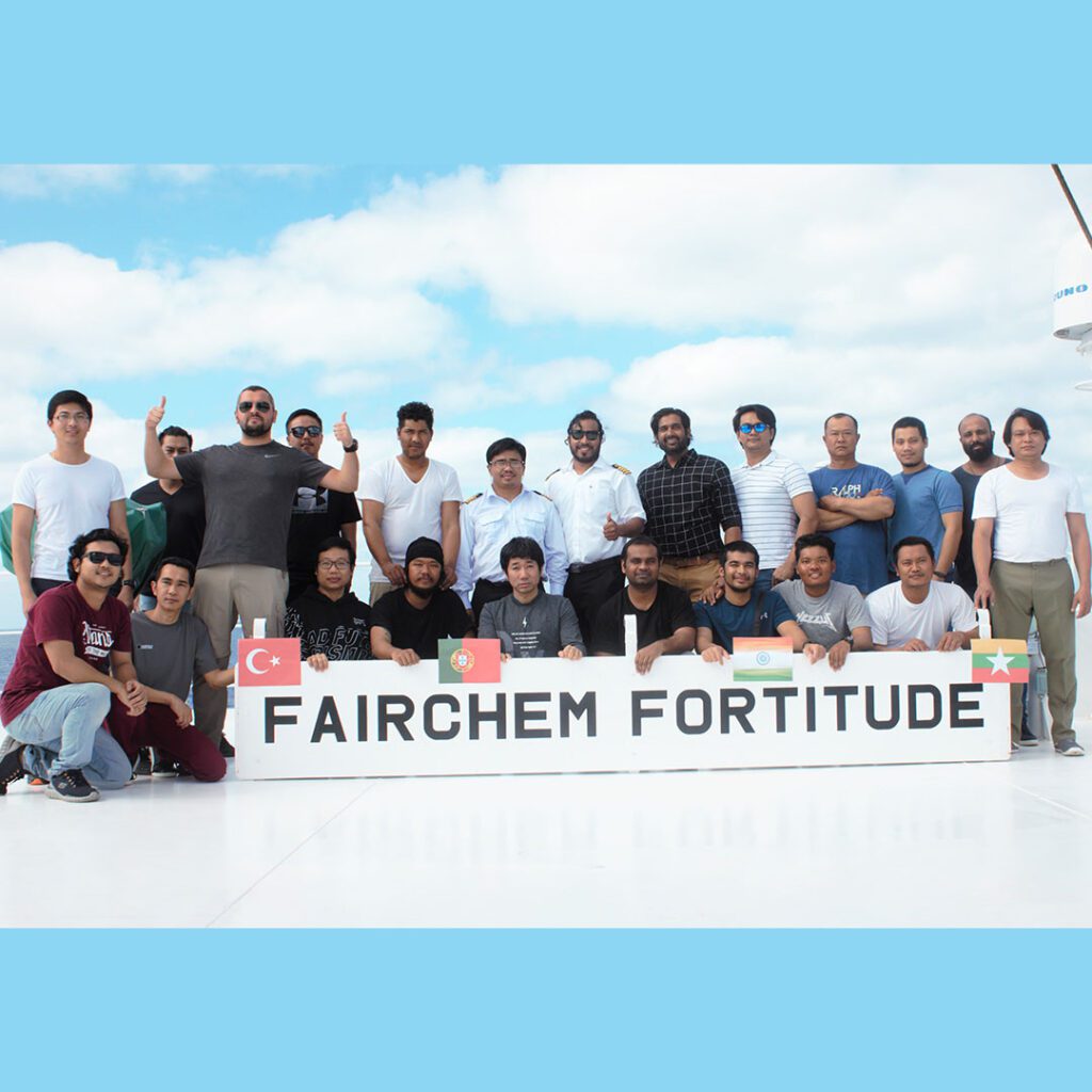 A diverse group of ship crew members posing together on the deck of the vessel "FAIRCHEM FORTITUDE" under a blue sky.