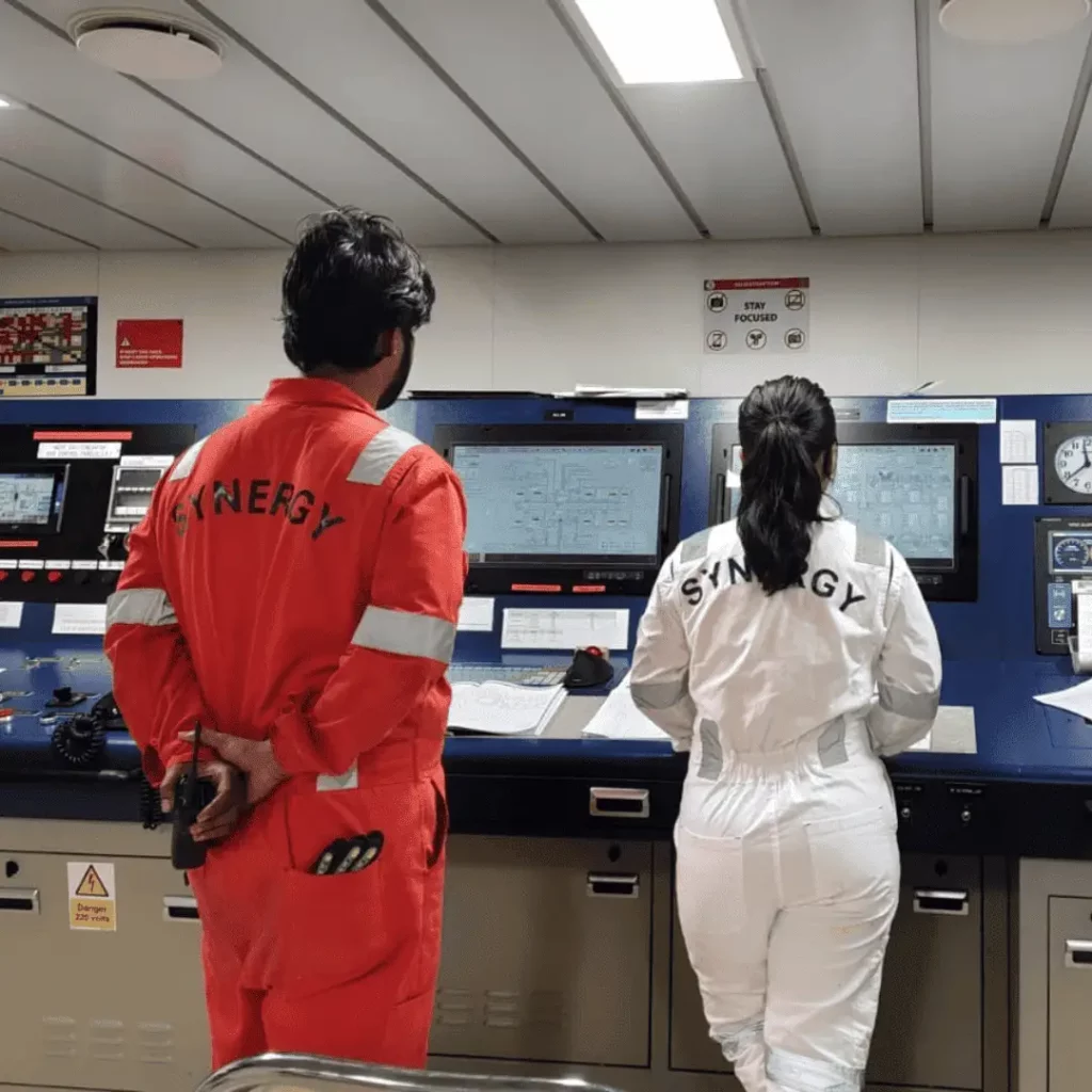 Synergy Marine Group engineers, wearing boiler suits, monitoring system screens inside the ship's engine control room.