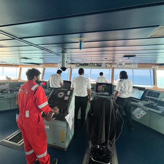 Able seaman steering at the helm wheel alongside ship officers on a vessel navigation bridge