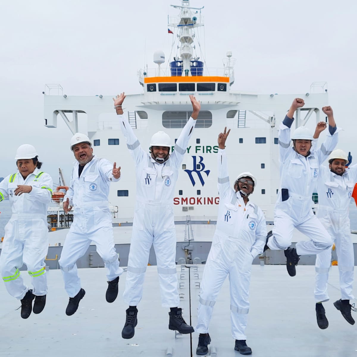 Group of seafarers in white coveralls and helmets jumping joyfully on a ship&rsquo;s deck, with the words &ldquo;Safety First&rdquo; and &ldquo;No Smoking&rdquo; visible on the ship&rsquo;s bridge