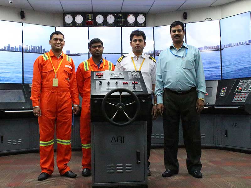 Four maritime professionals in uniform posing in a ship simulation room, standing behind a training helm labeled &ldquo;ARI.&rdquo;