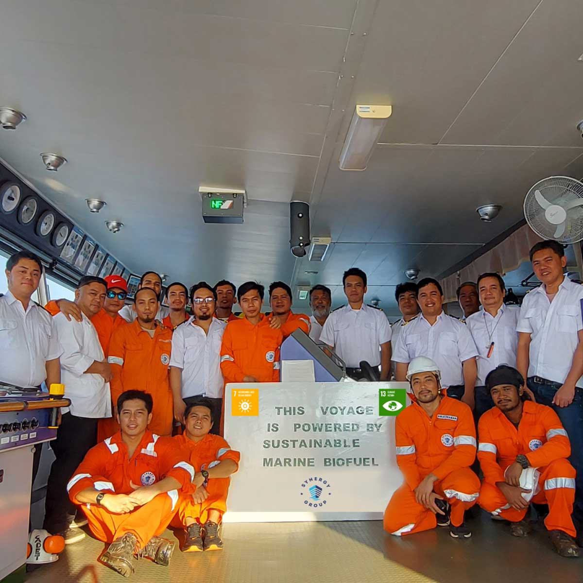Group of crew members in orange and white uniforms posing inside a ship&rsquo;s control room beside a sign that reads &ldquo;This voyage is powered by sustainable marine biofuel.&rdquo;