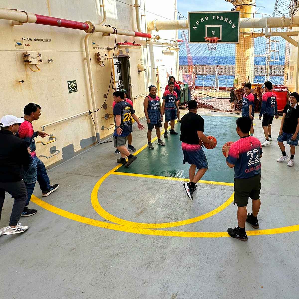 Crew members playing basketball on a ship&rsquo;s deck, wearing matching jerseys with &ldquo;Nord Ferrum&rdquo; printed on them, with the ocean visible in the background