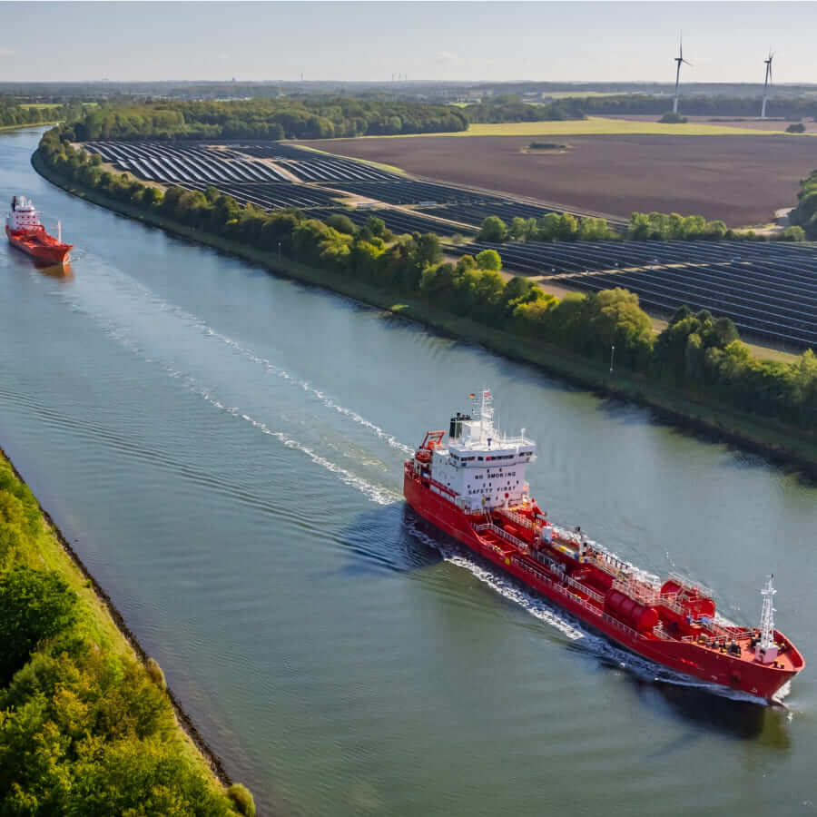 Red tankers sailing on a river with solar panels and wind turbines along the shore