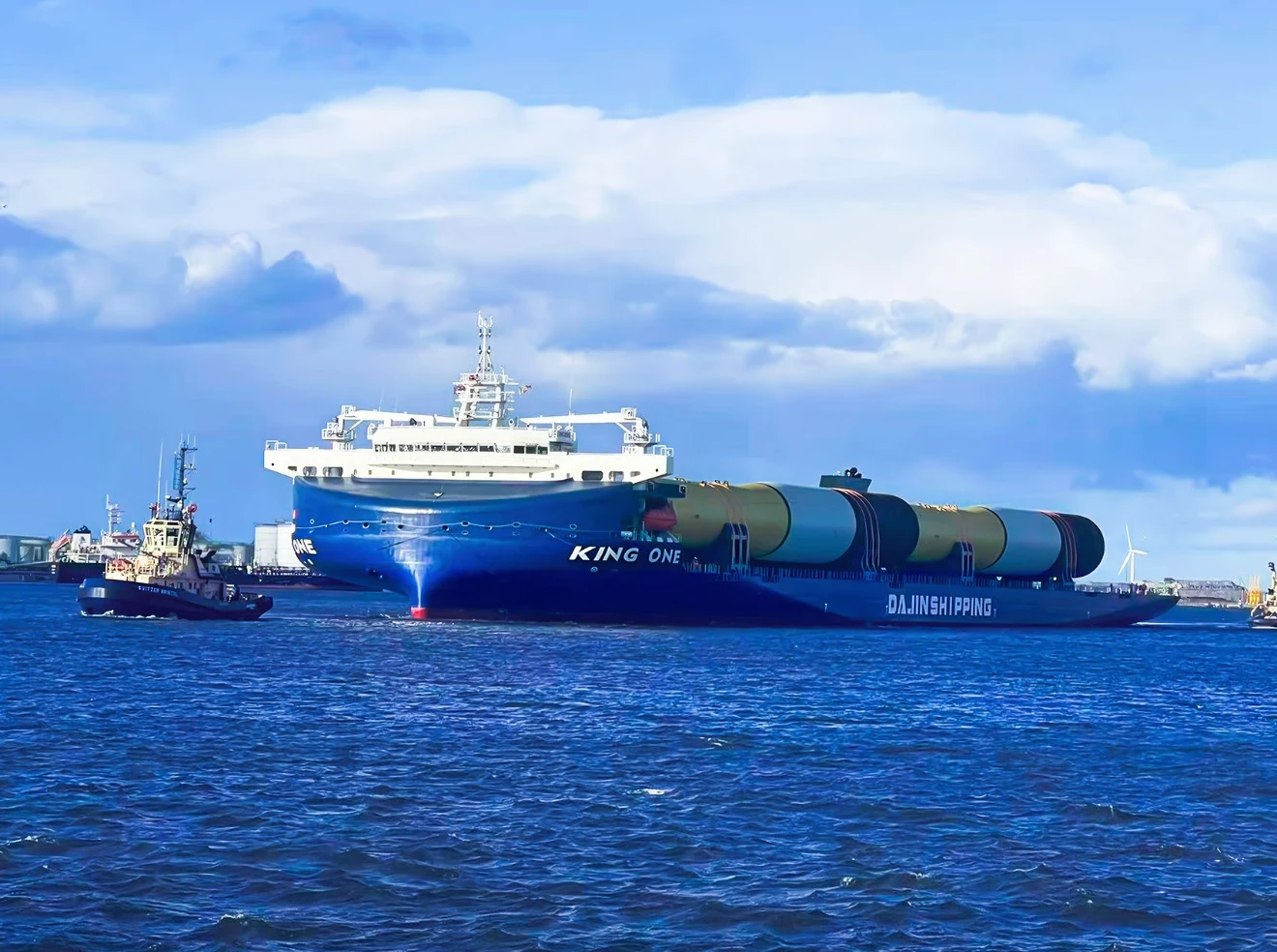 Heavy deck carrier KING ONE underway at sea, carrying multiple large cylindrical offshore wind monopiles securely fastened on its expansive open deck, with a tugboat assisting nearby and wind turbines visible in the distant background.