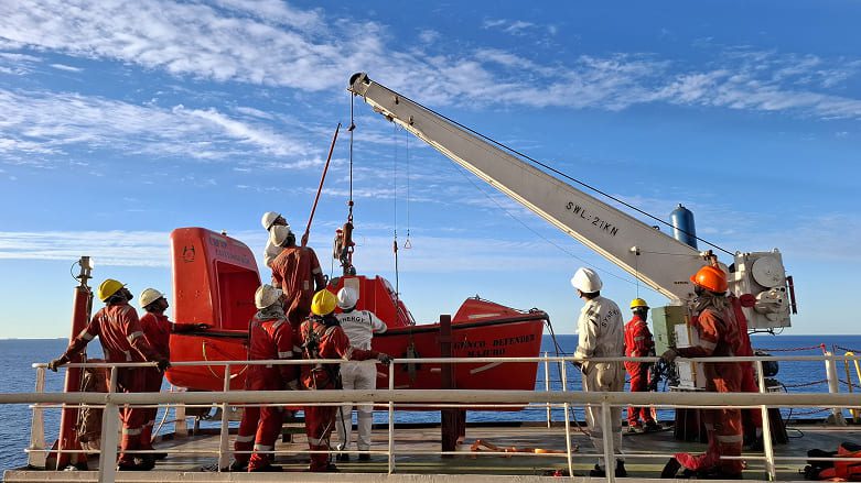 A well-composed lifeboat and davit scene in which the sweeping diagonal of the davit arm guides the eye toward a moment of teamwork, preparation and shipboard safety.